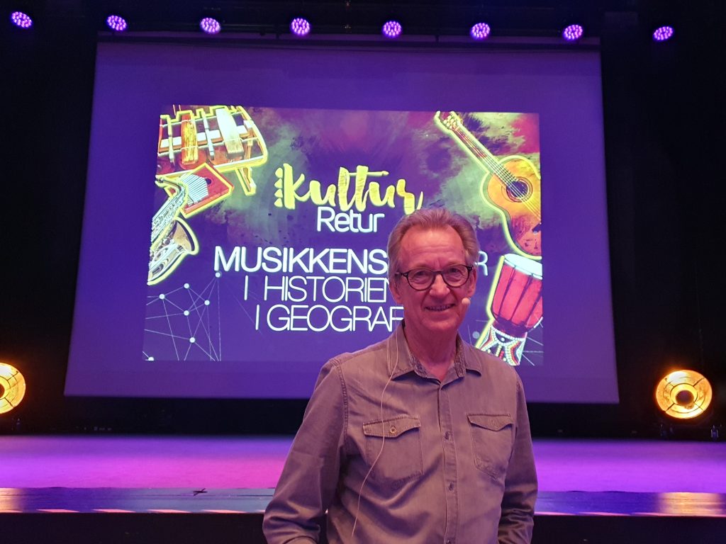 Fotografi av Sigbjørn Nedland. Han står foran en skjerm med teksten «Kultur retur, musikkens reiser i historien og geografien».