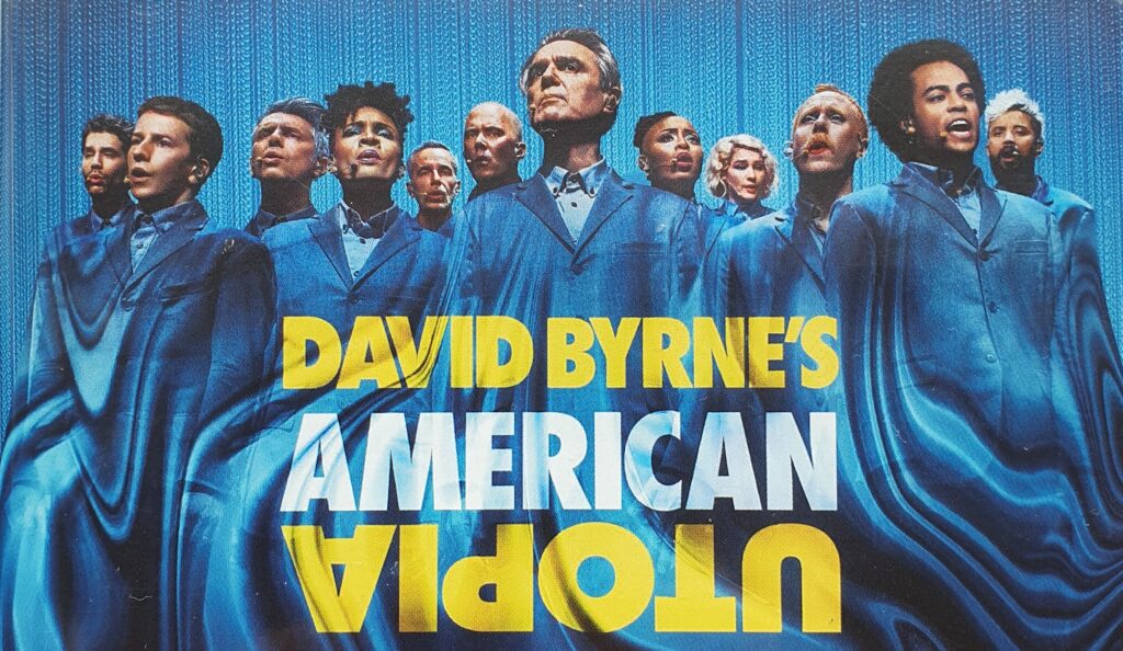 Tekst i bildet: David Byrne's American Utopia. Teksten står over flere mennesker kledd i blått.